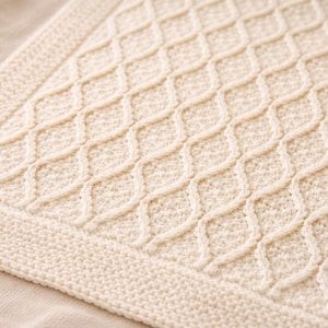 Könnte beinhalten: Cremefarbene Strickdecke mit sich wiederholendem Rautenmuster. Die Decke hat eine strukturierte Oberfläche und einen gestrickten Rand. Die weiche, neutrale Farbe und das komplizierte Design deuten auf einen gemütlichen, handgefertigten Artikel hin, der für die Inneneinrichtung geeignet ist.