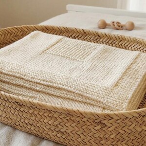 May include: A woven wicker basket with wooden handles holds a folded cream-colored knitted blanket. The blanket has a textured square design. A crib and other folded blankets are in the background.