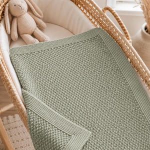 Può includere: Una coperta lavorata a maglia color verde salvia con un motivo testurizzato a grana di riso e un bordo liscio a legaccio. La coperta è drappeggiata su una culla in vimini intrecciato, accanto a un morbido coniglio di peluche beige chiaro.