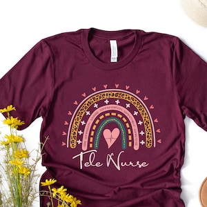 Könnte beinhalten: Ein maroonfarbenes T-Shirt mit einem Regenbogen-Design und dem Text "Tele Nurse". Der Regenbogen besteht aus verschiedenen Farben und Mustern, darunter Leopardenmuster, Herzen und Kreuze.