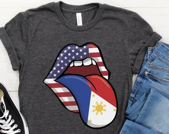 Puede incluir: Una camiseta gris oscuro con un gráfico de labios rojo, blanco y azul con una lengua que sobresale. La lengua es roja, blanca y azul con una estrella amarilla en el centro. El gráfico recuerda a las banderas estadounidense y filipina.