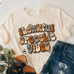 Può includere: Una maglietta beige con una grafica in stile retrò che dice "thankful speechie vibes" in lettere marroni, arancioni e grigie. La maglietta è abbinata a pantaloncini di jeans e sandali marroni.