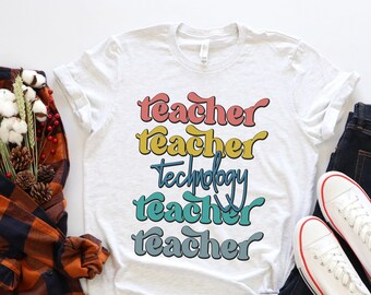 May include: A white t-shirt with the text "teacher teacher technology teacher teacher" in a rainbow of colors. The shirt is laid out on a white surface with a plaid shirt, a pair of jeans, and a pair of white sneakers.