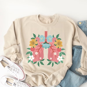 May include: A beige sweatshirt with a floral design featuring a stylized illustration of human lungs. The design includes pink, red, yellow, blue, and green flowers.
