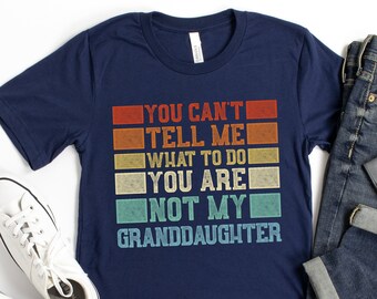 May include: Navy blue t-shirt with the text "YOU CAN'T TELL ME WHAT TO DO YOU ARE NOT MY GRANDDAUGHTER" in a retro color block design. The shirt is paired with blue jeans, a watch, and white sneakers.