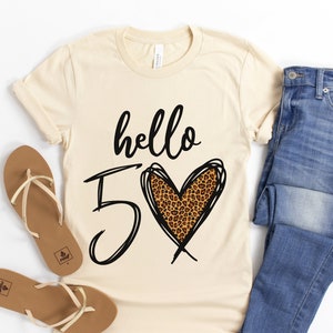 May include: A beige t-shirt with the text "hello 5" and a leopard print heart. The shirt is paired with blue jeans and brown sandals.