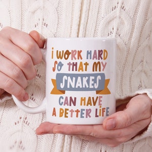 May include: White ceramic mug with a colorful design that reads "I work hard so that my snakes can have a better life."