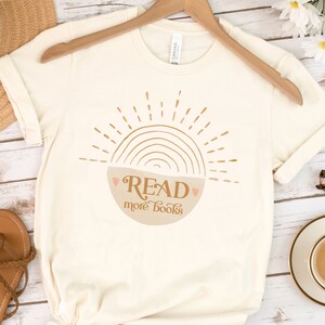 May include: A beige t-shirt with a graphic of a sun with a rainbow and a bowl that says "READ more books".