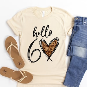 May include: Cream-colored t-shirt with the words "hello 60" in black script and a leopard print heart. The shirt is paired with blue jeans and brown sandals. The image is on a white background.