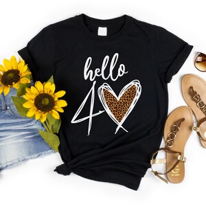Puede incluir: Camiseta negra con el texto "hello 40" y un diseño de corazón con estampado de leopardo. La camiseta se combina con pantalones cortos de mezclilla, gafas de sol, sandalias y girasoles. El logotipo de Down South Crafts está en la esquina superior izquierda.