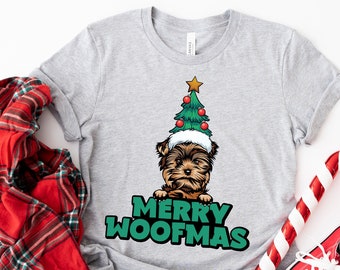 Może przedstawiać: Jasnoszara koszulka z Yorkshire Terrierem w czapce w kształcie choinki. Tekst "MERRY WOOFMAS" jest wydrukowany na zielono pod psem. Czerwono-biała laska cukrowa i czerwone trampki leżą obok koszulki.