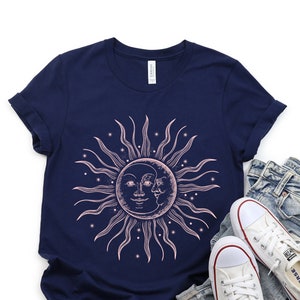 May include: A navy blue t-shirt with a pink sun and moon graphic. The sun has a face and the moon is partially visible behind the sun. The graphic is surrounded by small stars.