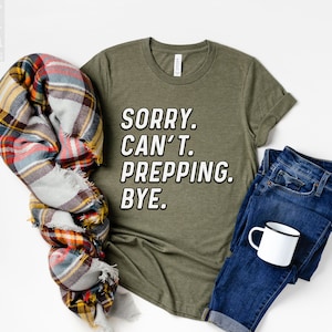 May include: A green t-shirt with the text "SORRY. CAN'T. PREPPING. BYE." printed in white on the front. The shirt is laid flat on a white surface with a plaid scarf, a pair of blue jeans, and a white mug.