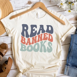 Puede incluir: Una camiseta blanca con el texto "READ BANNED BOOKS" en una fuente retro y colorida. La camiseta está en una percha y tiene un corte holgado.