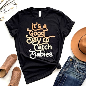 May include: Black t-shirt with a white, orange, and yellow graphic that says "It's a Good Day to Catch Babies".