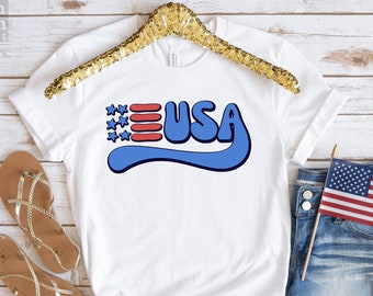 Può includere: Maglietta bianca con una grafica "USA" blu e rossa. La grafica presenta una bandiera americana stilizzata con stelle e strisce.