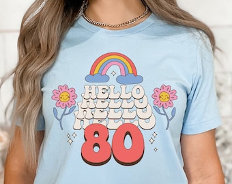 Può includere: Maglietta blu chiaro con un arcobaleno, fiori e il testo "HELLO 80" in stile retrò.
