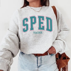 K&ouml;nnte beinhalten: Ein grauer Pullover mit dem Text "SPED TEACHER" in t&uuml;rkisfarbener und rosafarbener Schrift.
