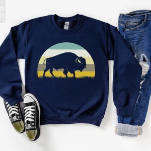 May include: Navy blue sweatshirt featuring a silhouette of a bison against a retro-style sunset design with horizontal stripes in teal, white, yellow, and gray. The sweatshirt is paired with blue jeans and black sneakers.