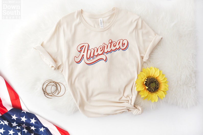 May include: A beige t-shirt with the word "America" printed in red, white, and blue retro lettering. The shirt is knotted at the bottom and is photographed on a white surface with a sunflower and an American flag.