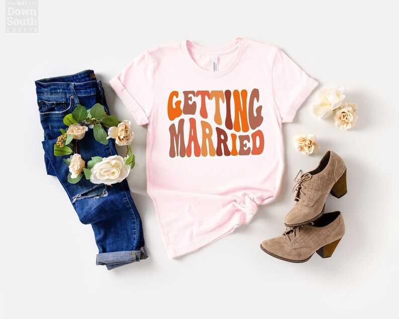 May include: A pink t-shirt with the text "GETTING MARRIED" in a retro, colorful font. The shirt is laid flat on a white background with a pair of brown suede shoes and a pair of blue jeans with a floral crown.