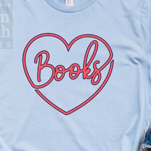 May include: A light blue t-shirt with a red heart outline and the word "Books" in red lettering inside the heart. The shirt is on a wooden surface with a pair of white sneakers and a pair of blue jeans.