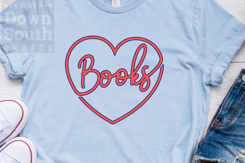 May include: A light blue t-shirt with a red heart outline and the word "Books" in red lettering inside the heart. The shirt is on a wooden surface with a pair of white sneakers and a pair of blue jeans.