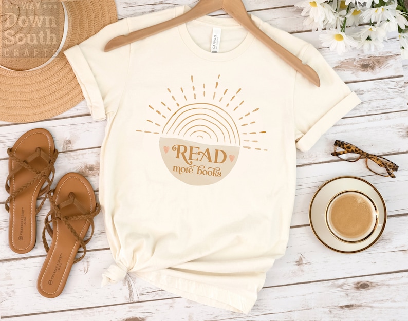 May include: A beige t-shirt with a graphic of a sun with a rainbow and a bowl that says "READ more books".
