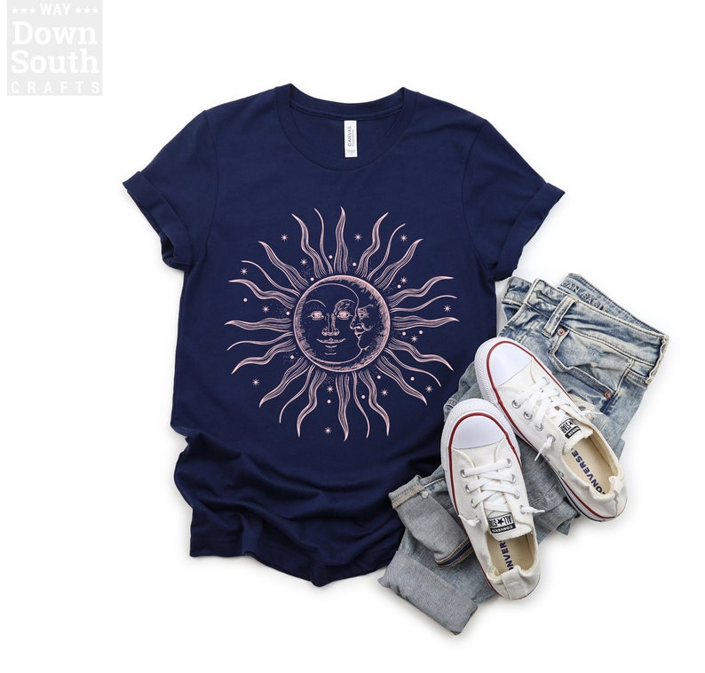 May include: A navy blue t-shirt with a pink sun and moon graphic. The sun has a face and the moon is partially visible behind the sun. The graphic is surrounded by small stars.