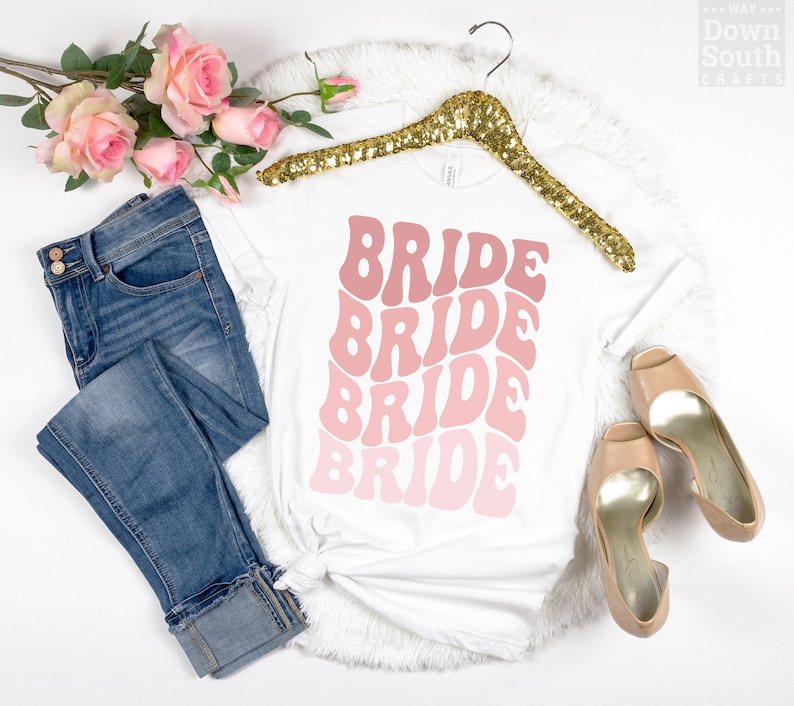 May include: White t-shirt with the word "BRIDE" repeated four times in a pink ombre design. The shirt is on a hanger with a pair of jeans and shoes.