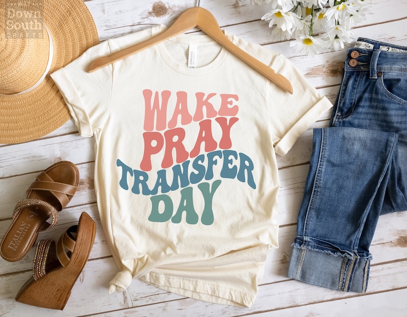 May include: A white t-shirt with the text "WAKE PRAY TRANSFER DAY" in a retro font with pink, red, blue, and green colors.