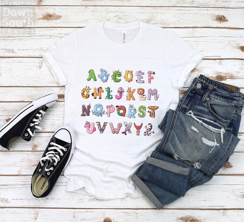May include: White t-shirt with a colorful alphabet printed on it. Each letter is decorated with a different animal or creature. The shirt is laid flat on a white wooden surface with a pair of black sneakers and blue jeans.