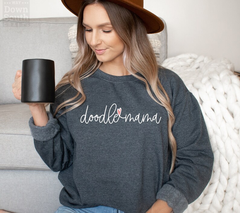 Pu&ograve; includere: Felpa grigio scuro con le parole "doodle mama" in corsivo bianco e un piccolo cuore rosa. La felpa ha un girocollo e maniche lunghe. Una tazza nera &egrave; tenuta nella mano sinistra.