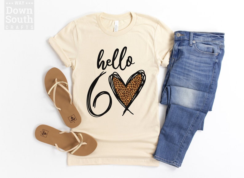 May include: Cream-colored t-shirt with the words "hello 60" in black script and a leopard print heart. The shirt is paired with blue jeans and brown sandals. The image is on a white background.