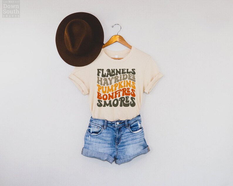 Puede incluir: Una camiseta beige con un dise&ntilde;o gr&aacute;fico que dice "Flannels, Hayrides, Pumpkins, Bonfires, Smores" en una fuente retro con colores naranja, amarillo y marr&oacute;n.