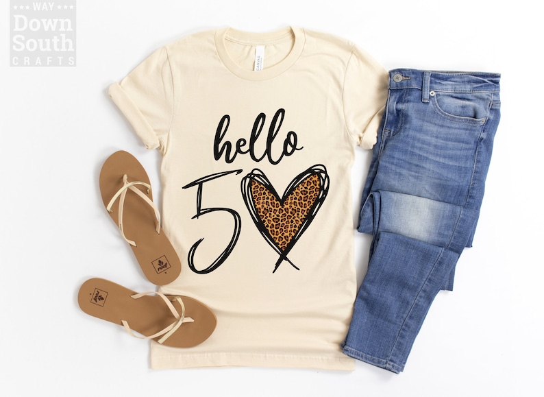 May include: A beige t-shirt with the text "hello 5" and a leopard print heart. The shirt is paired with blue jeans and brown sandals.