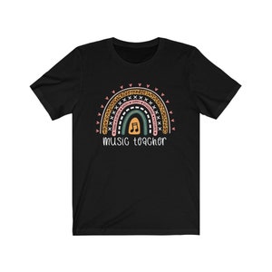 以下が含まれることがあります： 虹のデザインが施された黒いTシャツ。虹には「Today, Inspire, Teach, Sing, Learn, Encourage, Positive」という文字と音符が描かれている。「music teacher」という文字が虹の下にある。