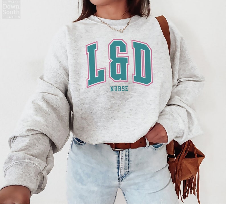 Puede incluir: Una sudadera gris con el texto "L&D Nurse" en letras turquesa y rosa. La sudadera tiene un corte holgado y es perfecta para el uso casual.