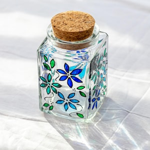 May include: A clear, square glass jar with a cork stopper. The jar is decorated with blue and green floral designs. The jar is sitting on a white surface.