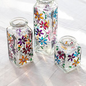 May include: Three clear glass vases of different sizes, decorated with vibrant floral patterns. The flowers are painted in red, orange, purple, blue, and yellow, with green leaves. The square-shaped vases are displayed on a white surface.