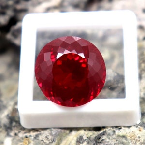 Puede incluir: Una gema roja vibrante, probablemente un rubí, se muestra en una caja cuadrada blanca. La caja está sentada sobre una superficie rugosa y texturizada.