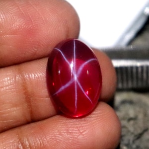 May include: A polished, oval-shaped ruby gemstone with a six-rayed star pattern. The gem is a deep red color with white star lines. The ruby is held in a hand, with a blurred background.