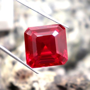 Puede incluir: Una piedra preciosa roja oscura con un corte cuadrado esmeralda. La piedra preciosa está sujeta por unas pinzas.