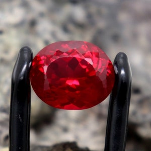 Puede incluir: Una piedra preciosa ovalada de color rojo intenso con un corte facetado, sujeta en una abrazadera de metal negro.