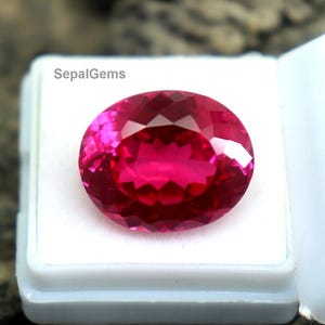 Puede incluir: Una gema rosa intenso, tallada en forma ovalada, exhibida en una caja de plástico blanco. La gema tiene un corte brillante, reflejando la luz. La caja está abierta, revelando la gema sobre un fondo texturizado gris-marrón. El texto "SepalGems" es visible.