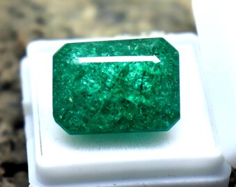 Könnte beinhalten: Ein lebendiger grüner Smaragd-Edelstein mit rechteckigem Schliff, präsentiert in einer offenen weißen Kunststoffbox. Der Edelstein hat eine satte, tiefe Farbe und ist vor einem strukturierten, dunkelbraunen Hintergrund platziert.