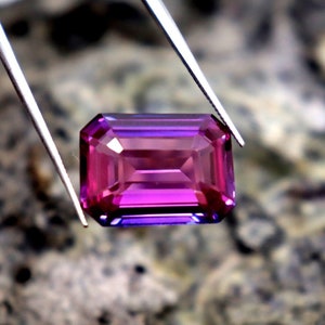 May include: A vibrant purple and blue gemstone with a rectangular emerald cut. The gemstone is held by tweezers against a blurred background.