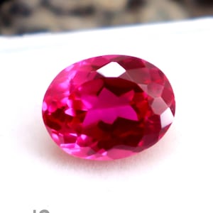 Peut inclure: Une pierre précieuse rouge vif, de forme ovale et taillée à facettes, présentée dans un écrin en plastique blanc. La gemme est brillante et réfléchit la lumière. Le texte "SepalGem" est imprimé sur le boîtier. Fond flou.