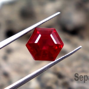 May include: A faceted, hexagonal, deep red gemstone is held by silver tweezers against a blurred background. The gem has a polished surface and reflects light, showcasing its facets. The background is a textured, neutral colour.