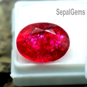 Puede incluir: Una vibrante gema roja, de corte ovalado, se exhibe en una caja de plástico blanco. La gema tiene una superficie facetada que refleja la luz, mostrando su rico color. El texto "SepalGems" es visible.